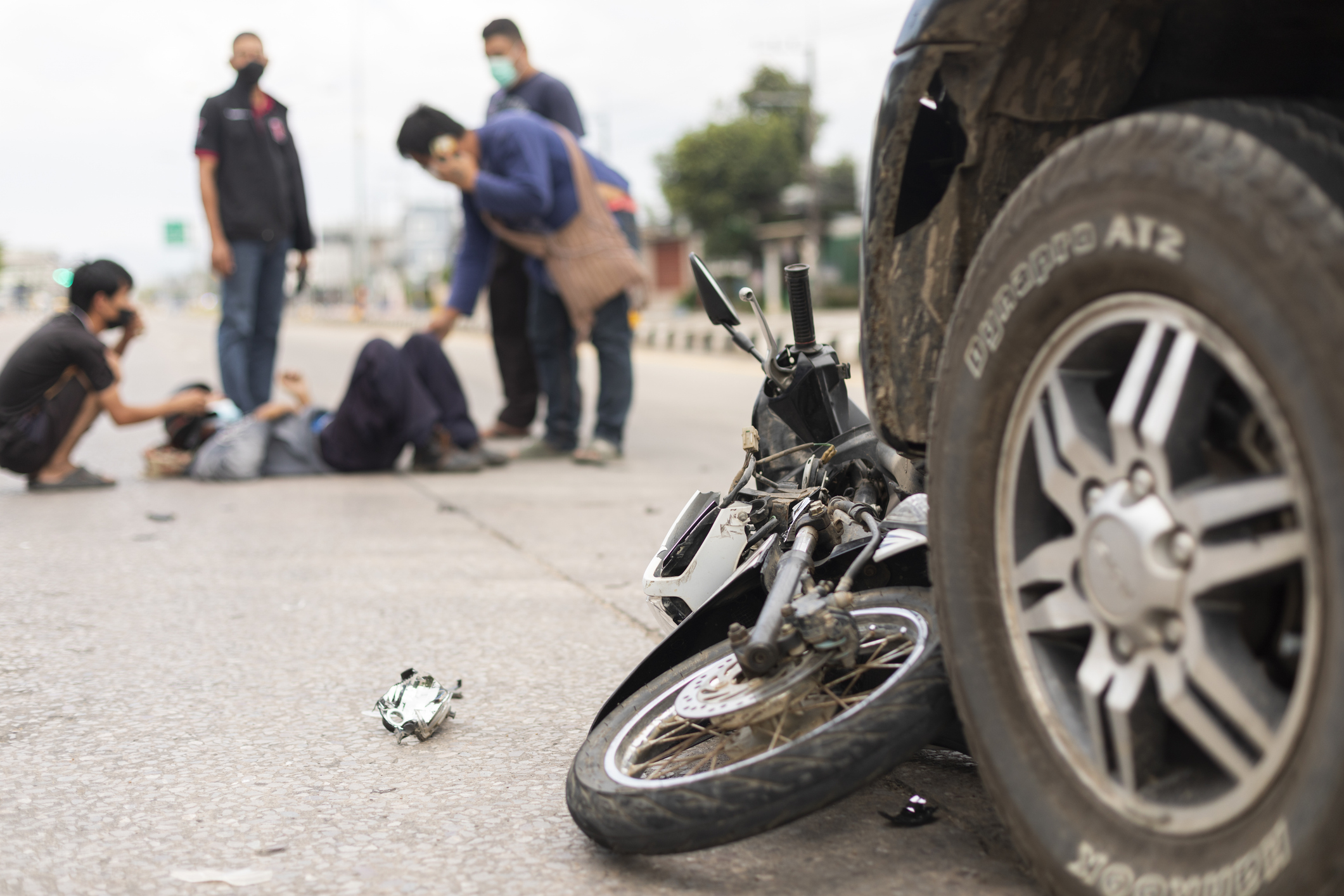 Car wheel with motorcycle underneath and group of people surrounding biker on ground in background